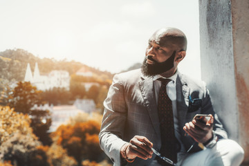 A handsome mature bearded African man in a formal suit with a cellphone and eyeglasses in his hands; an adult elegant bald black guy with a beard is using his smartphone outdoors, landscape behind him - Powered by Adobe