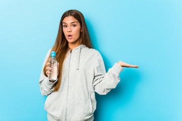 Young fitness woman holding a water bottle impressed holding copy space on palm.