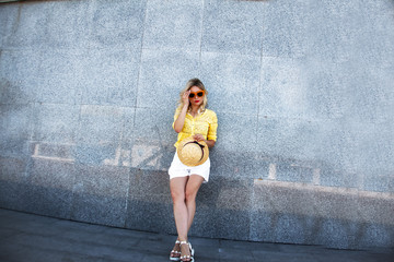 Pretty female model posing against the gray wall. Woman dressed urban style clothes , white shorts, yellow t-shirt and straw hat. The fashion street lifestyle of summer vacation.