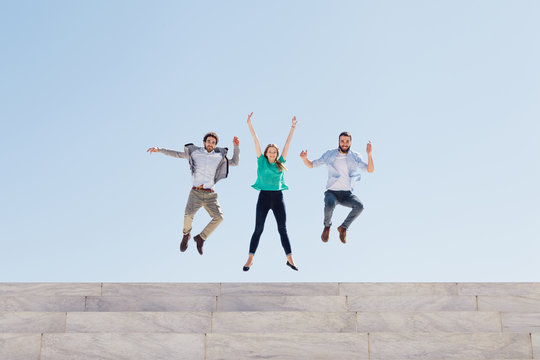Three friends jump into the air at the top of an external staircase of a building, behind them the blue sky of a spring day - Copy space - Millennial have fun together