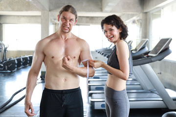Fototapeta na wymiar Young sporty and healthy caucasian woman wearing sportswear using measuring tape to handsome caucasian muscle man in gym with smiling face