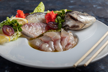 Light-salted dorado with sunflower in truffle oil. Next to slices of lemon, lime, grapes, pickled ginger. To the side of the plate are chopsticks.