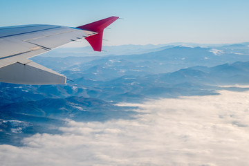 Fototapeta na wymiar Above the clouds. Wing aircraft in altitude during flight, alternating mountain ranges partially covered with thick white clouds, clear blue sky in the bckground. Flight over the Balkans.