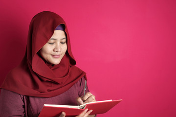 Happy confident successful Asian muslim woman wearing hijab  writing on book, smiling and thinking