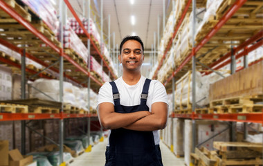logistic business and people concept - happy smiling indian loader or worker with crossed arms over warehouse background