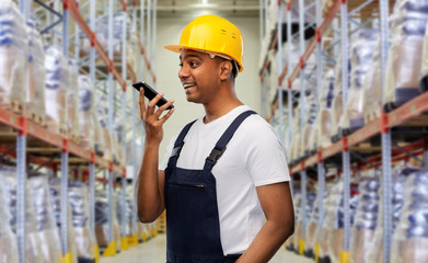 logistic business, technology and people concept - happy smiling indian loader or worker in helmet using voice command recorder on smartphone over warehouse background