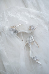 Wedding shoes with rhinestones for the bride who lie on a white veil fabric next to the bride's dress.