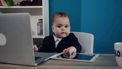 Cute baby sits at the table in formal wear as a baby boss, plays businessman at office, concentrated at work. Cuteness, humor, having fun. Positive emotions, success, joy of life. Child portrait