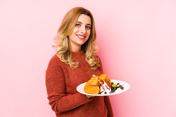 Young blonde woman eating a waffle dessert isolated looks aside smiling, cheerful and pleasant.