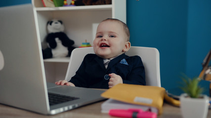 Adorable little toddler cutie in business suit happily smiling while sitting by the table in front of the computer. Baby boss concept. Successful lifestyle, happy childhood, having fun. Child’s