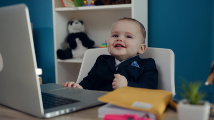 Adorable little toddler cutie in business suit happily smiling while sitting by the table in front of the computer. Baby boss concept. Successful lifestyle, happy childhood, having fun. Child’s