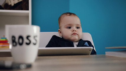 Selective focus on boss’s mug on the table, then on cute adorable baby boy in formal costume holding tablet. Successful childhood, happiness, serious baby. Early childhood education. Close up view.