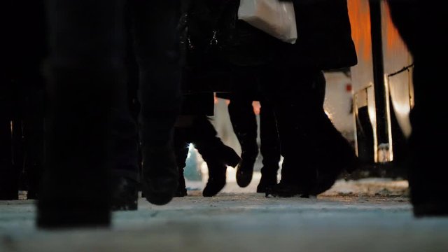 Crowd of pedestrians walking on tram or bus stop in blurred glare bokeh background. Feet of people walking busy city street at night during winter. Slow motion.