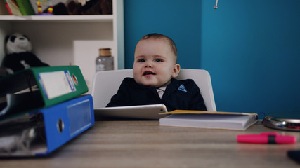 Fototapeta na wymiar Extremely cute toddler baby boss sits in the office, unrecognizable hand puts in front of the baby a pile of documents, baby boy sadly hilariously looks at it. Successful child, lots of work, working