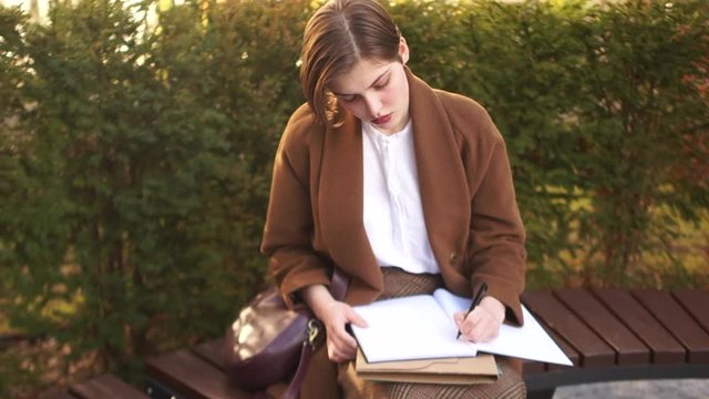 Short-haired girl sits on a bench in a park and writes something in a large notebook. Student preparing for the exam