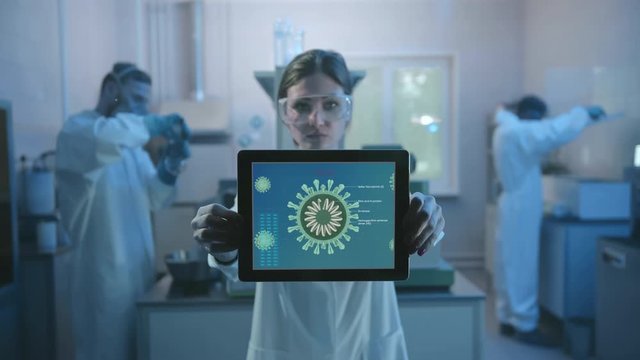 Doctor or researcher holds tablet PC with coronavirus illustration image to camera, standing in modern laboratory. Woman in protective antiplague suit on a background of scientits working