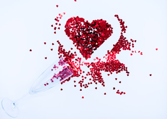 Champagne glass with red sequins scattered in the form of a heart on a white backdrop. Background for Valentine's day. Top view. Flat lay.