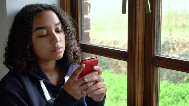 Sad beautiful mixed race African American girl teenager young woman using her mobile cell phone for social media or texting wearing a blue hoodie sitting looking out of a window 