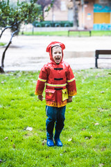 Funny child dressed in red raincoat firefighter play in a park on a rainy day.