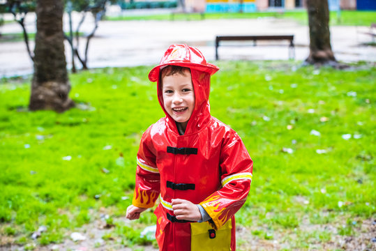Funny child dressed in red raincoat firefighter play in a park on a rainy day.