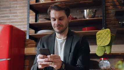 Young attractive male looking good and smiling with cheerful expression while texting his friends, using mobile phone, isolated on kitchen background. Technology, modern communication. Male portrait