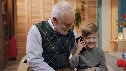 Old carrying grandfather looking for little boy in gray sweater. Nice child holding smartphone in hands and calling mom, laughing, listening to voice, speaking. Inside.