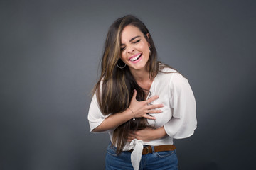 Joyful European student keeps hands crossed, laughs at good joke, wears casual clothes and round spectacles, standing indoors. Happy young woman with long hair poses against gray wall.