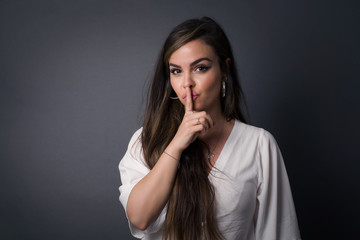 Dont tell my secret or not speak too loud, please! Emotional surprised student makes hush gesture, asks be quiet, has scared expression as afraids of revealing secret, poses against gray studio wall.