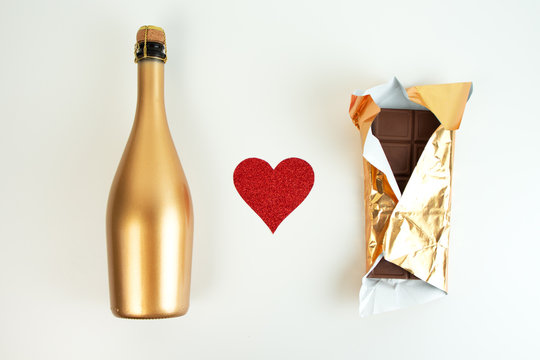 Golden champagne bottle and chocolate bar in gold wrapper and red glitter heart on a white background. Festive set. Top view