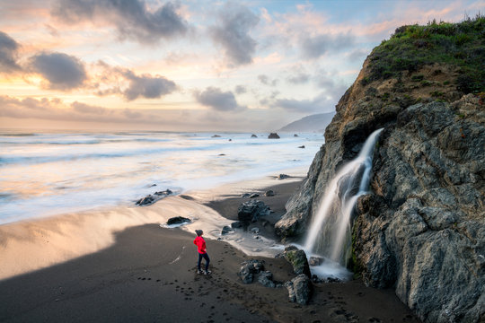 A woman stands on a black sand beach looking at a beautiful waterfall flowing into the ocean near the town of Westport in Mendocino County, California.