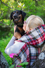  Blond woman with hood, holding her dog in arms in green and blooming nature. 