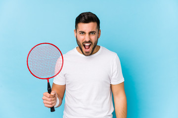 Young handsome man playing badminton isolated screaming very angry and aggressive.