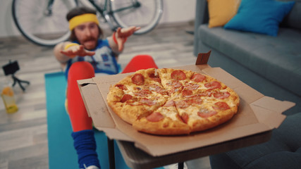 Funny fitness man doing abdominal crunches exercise failing to reach tasty hot pizza in the living...