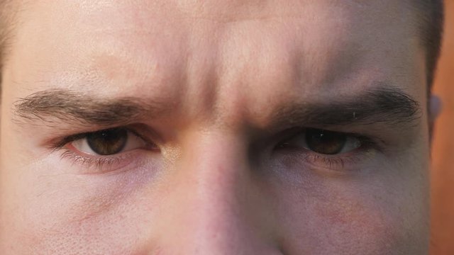 Close up brown eyes of guy staring and blinking with angry sight. Portrait of male face seriously looking into camera with negative emotion. Facial expression of confident young man