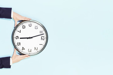 Hands of woman in business suit hold big plain wall clock on pastel blue background. six oclock....