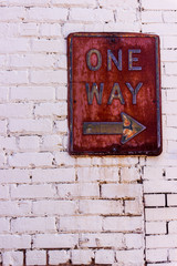 A rusty one way sign is attached to an old, white brick wall.
