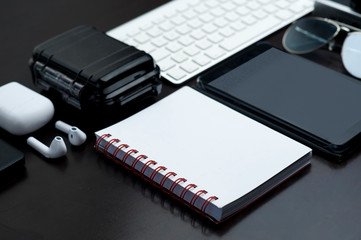 office table, with technology, white notebook, with red detail, headphones, waterproof case, computer keyboard, black tablet and sunglasses, black background