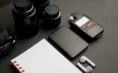 partial view work table photographer, with white notebook with red detail, black glasses, wireless headphones, two wafers, hard disk, and photometer and black waterproof case, on black background