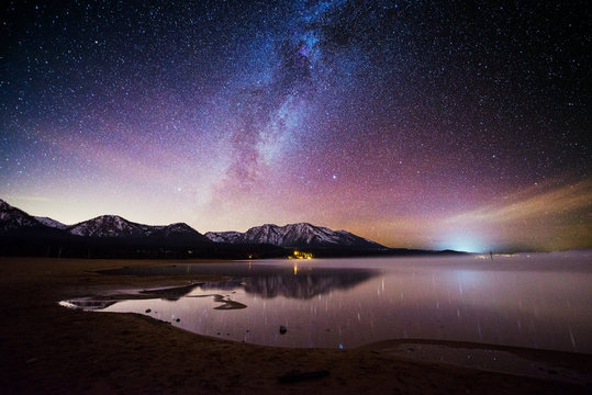 The Milky Way and a starry sky are illuminated above snow-capped mountains and Lake Tahoe at Kiva Beach, South Lake Tahoe, California