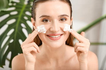 Beautiful young woman applying facial cream at home