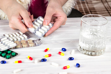 Close-up of bright colored medical pills and capsules with medicines in a woman's hand and a glass of water next to them. The woman takes the pills out of the blister.
