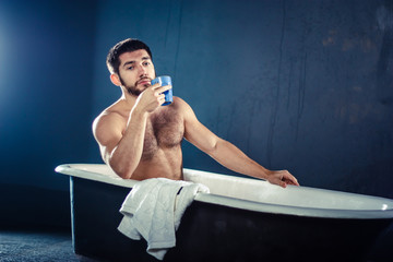 Handsome naked muscular adult bearded man taking bath and drinking coffee or tea, isolated on dark-blue background (with copyspace for your text) - obrazy, fototapety, plakaty