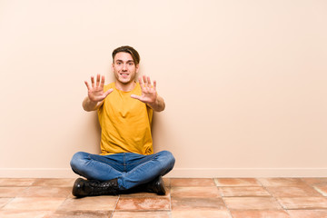 Young caucasian man sitting on the floor isolated showing number ten with hands.