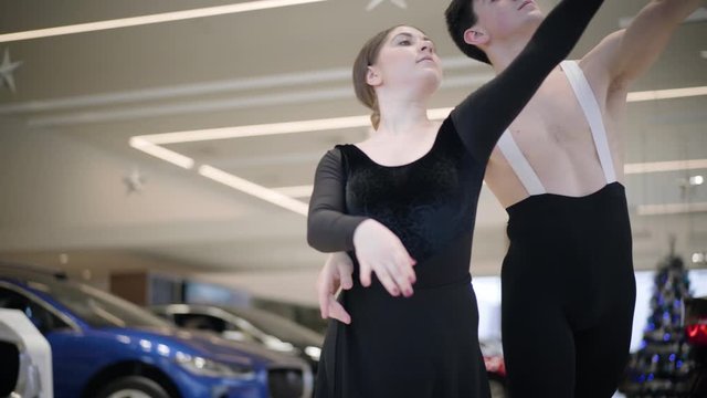 Camera following young Caucasian ballet dancers dancing in car dealership. Handsome man and beautiful woman performing classic dance in showroom. Art, elegance, automobile industry.