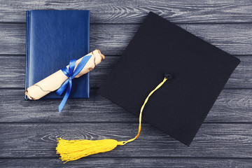 Graduation cap with diploma and notepad on black wooden table