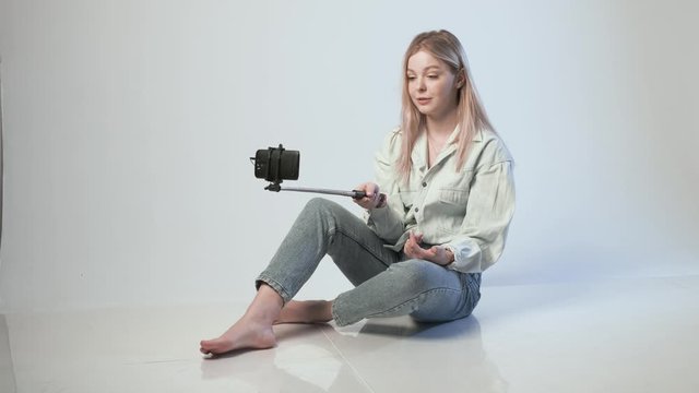 happy female records video with smartphone attached to selfie stick. blond girl filming vlog sitting on floor, smiling, laughing, using cellphone on white background. Beauty blogger recording blog