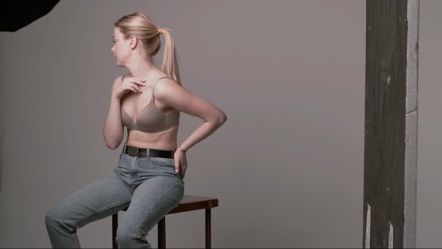 backstage of caucasian woman with long hair posing in beige lingerie and blue jeans on white studio background. model tests of pretty girl in bra. young female sitting on chair. body positive concept