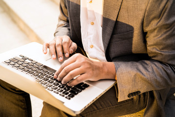 Young mixed race man in a white shirt and a business suit is sitting on the steps with a laptop and typing. Business lunch happy freelancer programmer. Break time outdoor. Hands close up