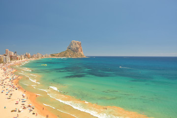 Colorful Mediterranean seascape.People sunbathe on the on the sea coast under umbrellas. In the distance, high-rise buildings and hotels of the resort town of Calpe and Mountain Penyal d'Ifach. Spain.
