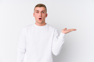 Young caucasian man on white background impressed holding copy space on palm.